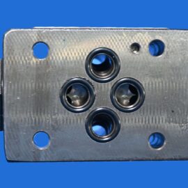 Close-up of a metal valve block with five circular ports and a central bore, set against a bright blue background.